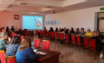 Sala konferencyjna. Przy półokragłych stołach siedzą ludzie. Na ścianie wyświetlana jest prezentacia multimedialna. Z prawej strony na ścianie wiszą tablice oraz znajdują się szklane drzwi. Z lewej strony widać częściowo mównicę oraz stojak z flagami. 